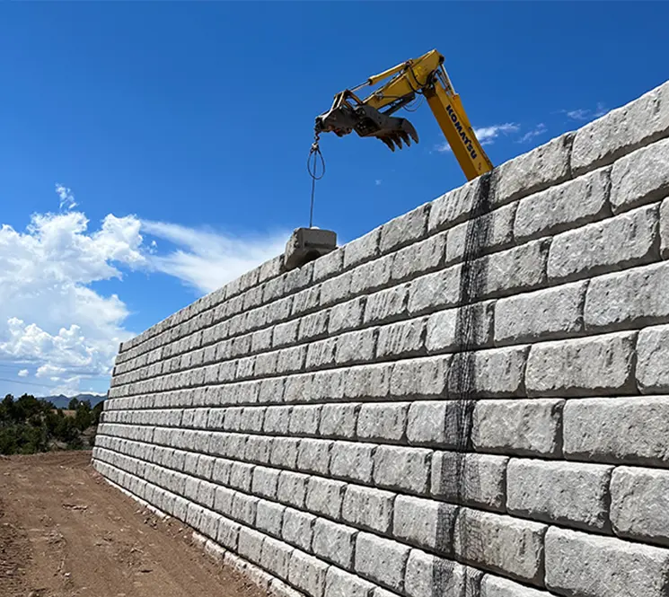 A crane lifting a large concrete block to place it on top of a massive retaining wall under a sunny sky.