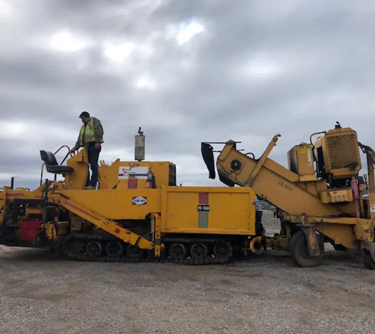 Worker operating a yellow asphalt paving machine under a cloudy sky.