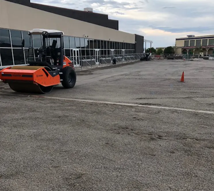 Large roller compactor smoothing a parking lot surface near a commercial building.