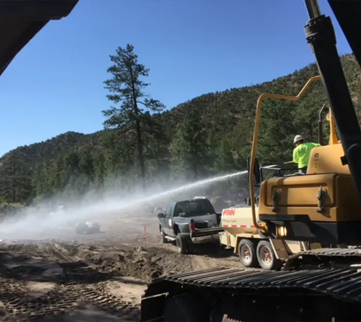 A construction worker operating a water truck, spraying water on a dusty road at a site in a mountainous area.