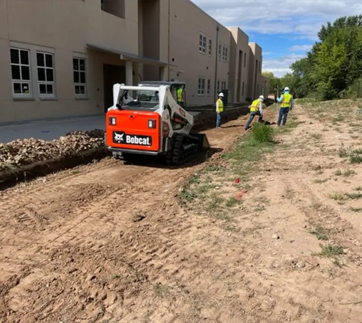 A Bobcat skid-steer loader grading a dirt path near a commercial building while workers in safety vests stand nearby.