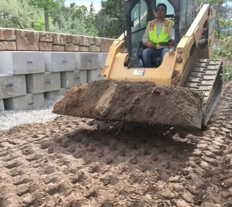 A construction worker operating a Caterpillar skid-steer loader, moving dirt near a retaining wall made of large stone blocks.