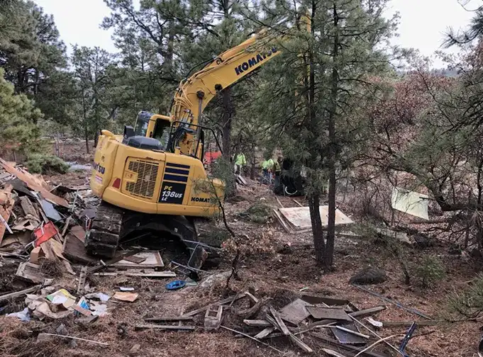 Komatsu excavator demolishing and clearing debris from a wooded site with construction workers in the background.