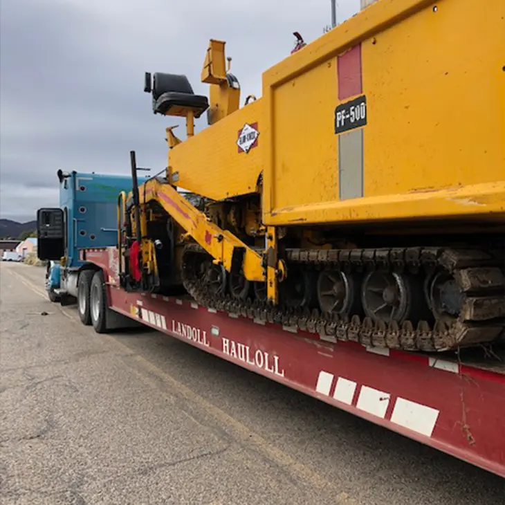 Yellow paving machine loaded onto a red Landoll trailer, secured for transport by a blue semi-truck.