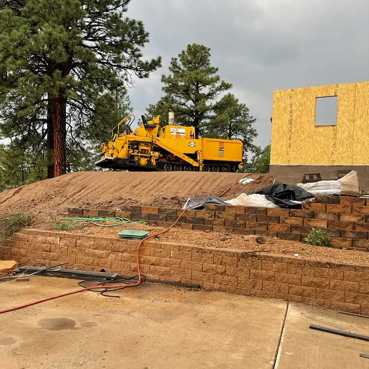 Yellow paving machine parked on a leveled dirt surface near a construction site, with retaining walls and an unfinished building in the foreground.