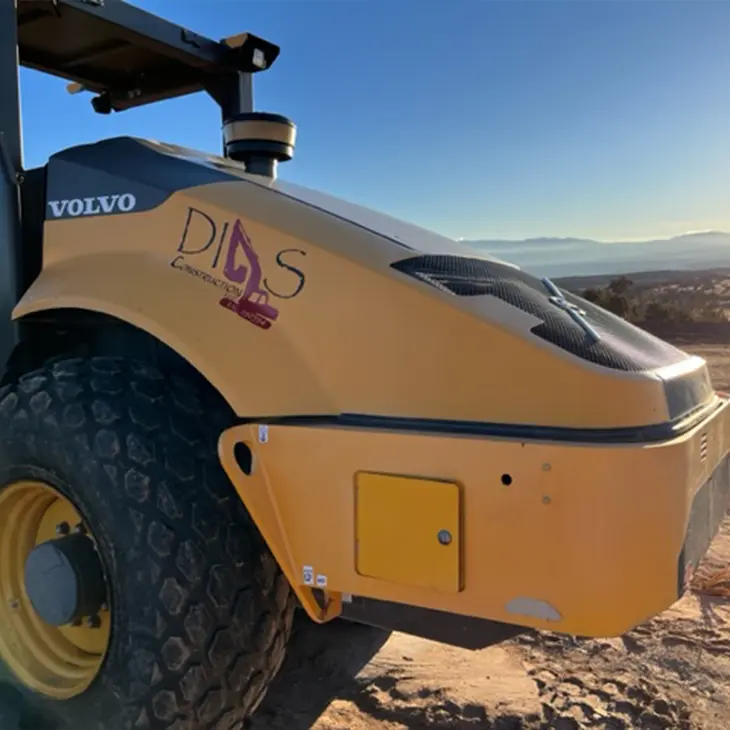Close-up of a yellow Volvo construction compactor with the Digs Construction logo, parked on a dirt site with mountains and a sunset in the background.